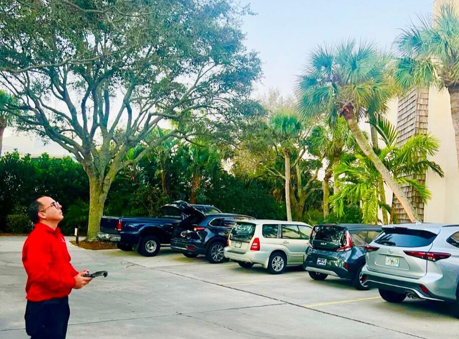 A person in a red shirt operating a flying drone in a parking lot with trees and cars.