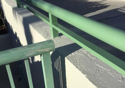 A sunlit green railing casting a shadow on a concrete surface near tropical foliage.