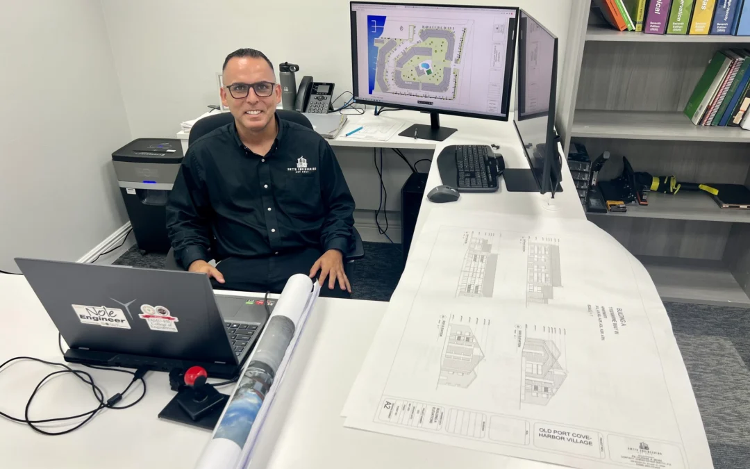 Person at a desk with a laptop, dual monitors displaying plans, and blueprints on the table.