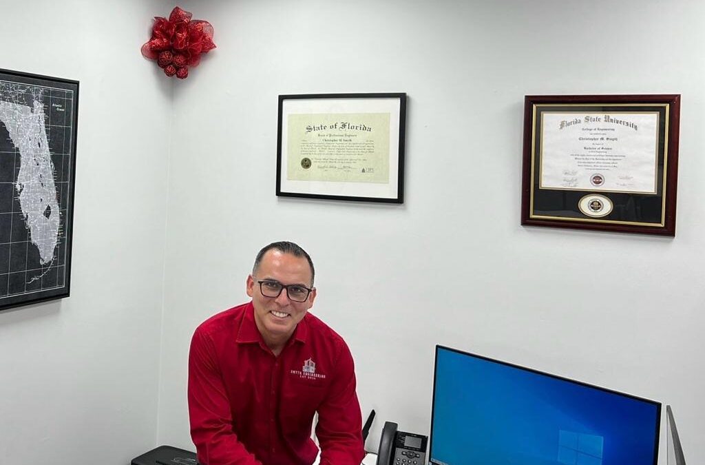 Person in red shirt sitting by a desk with a computer, phone, and diplomas on the wall.