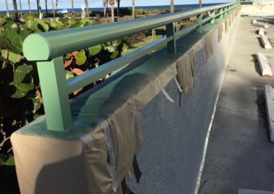 Seaside walkway with a green railing wrapped in protective paper, flanked by palm trees under a blue sky.